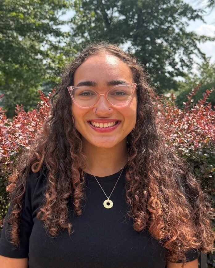 Young woman with curly hair and pink glasses smiling outdoors, wearing a black top and a circular pendant necklace.