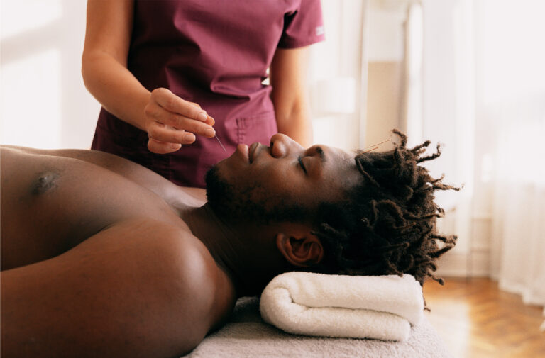 Shirtless man lying on a massage table with a towel under his head while a therapist prepares to treat his chest area.